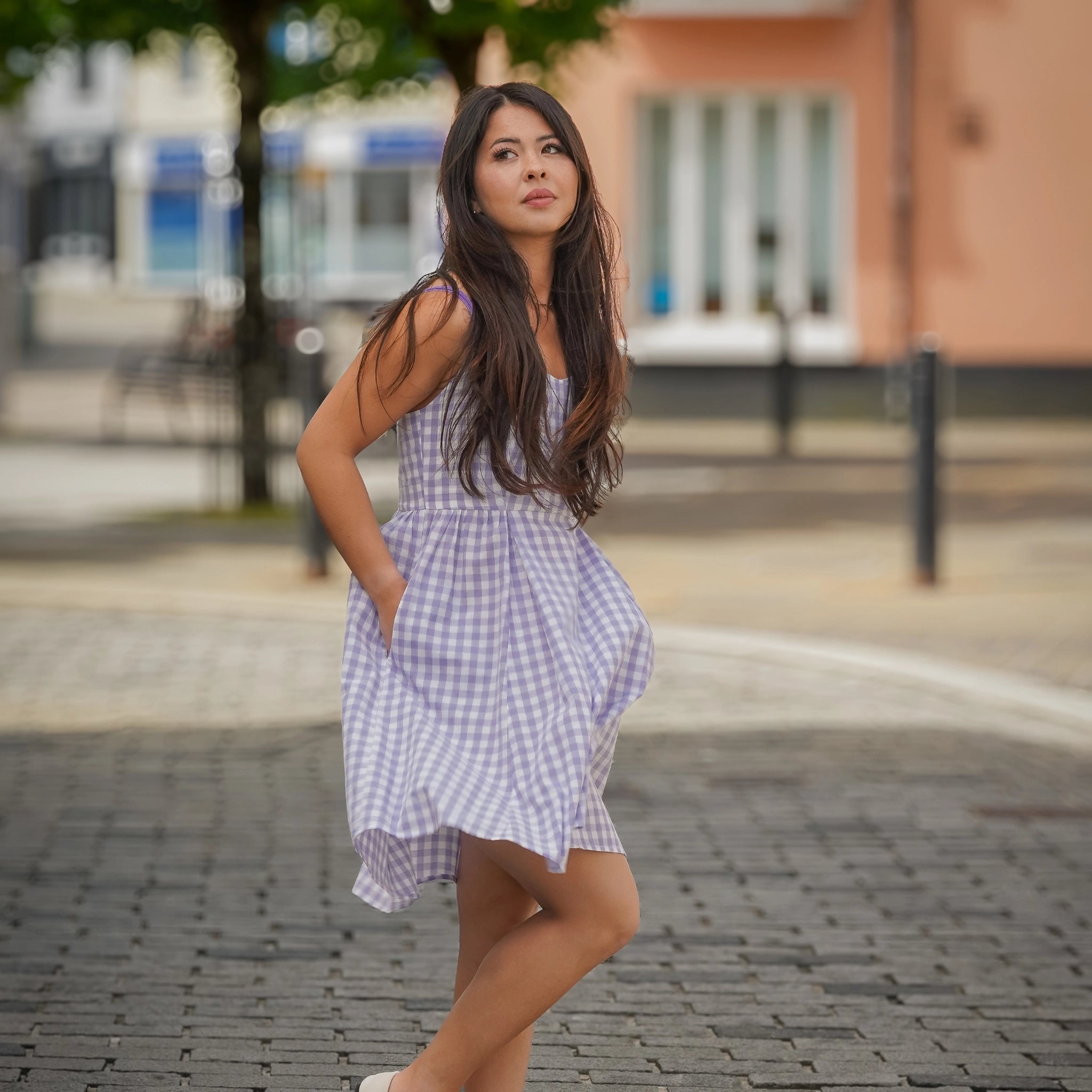 A woman in a checkered dress stands with her hands in her pockets on a cobblestone street, with buildings and a tree in the background.