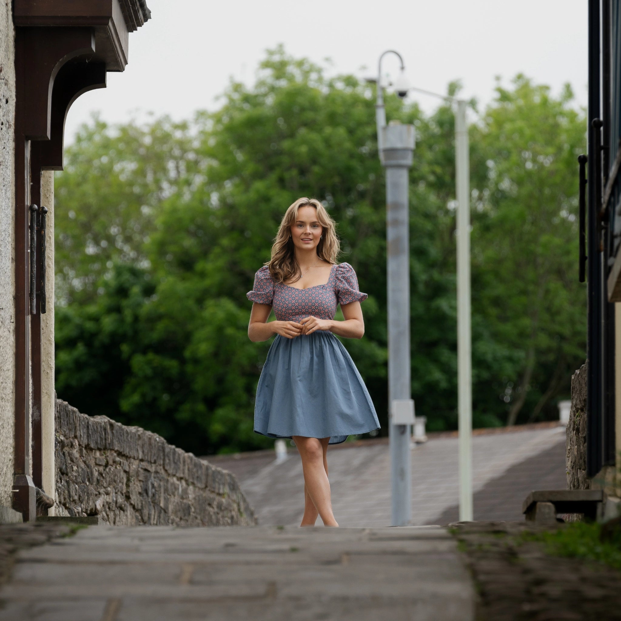 A woman stands outdoors on a stone walkway, framed by buildings and greenery in the background. She is wearing a blue skirt and a light-colored top.