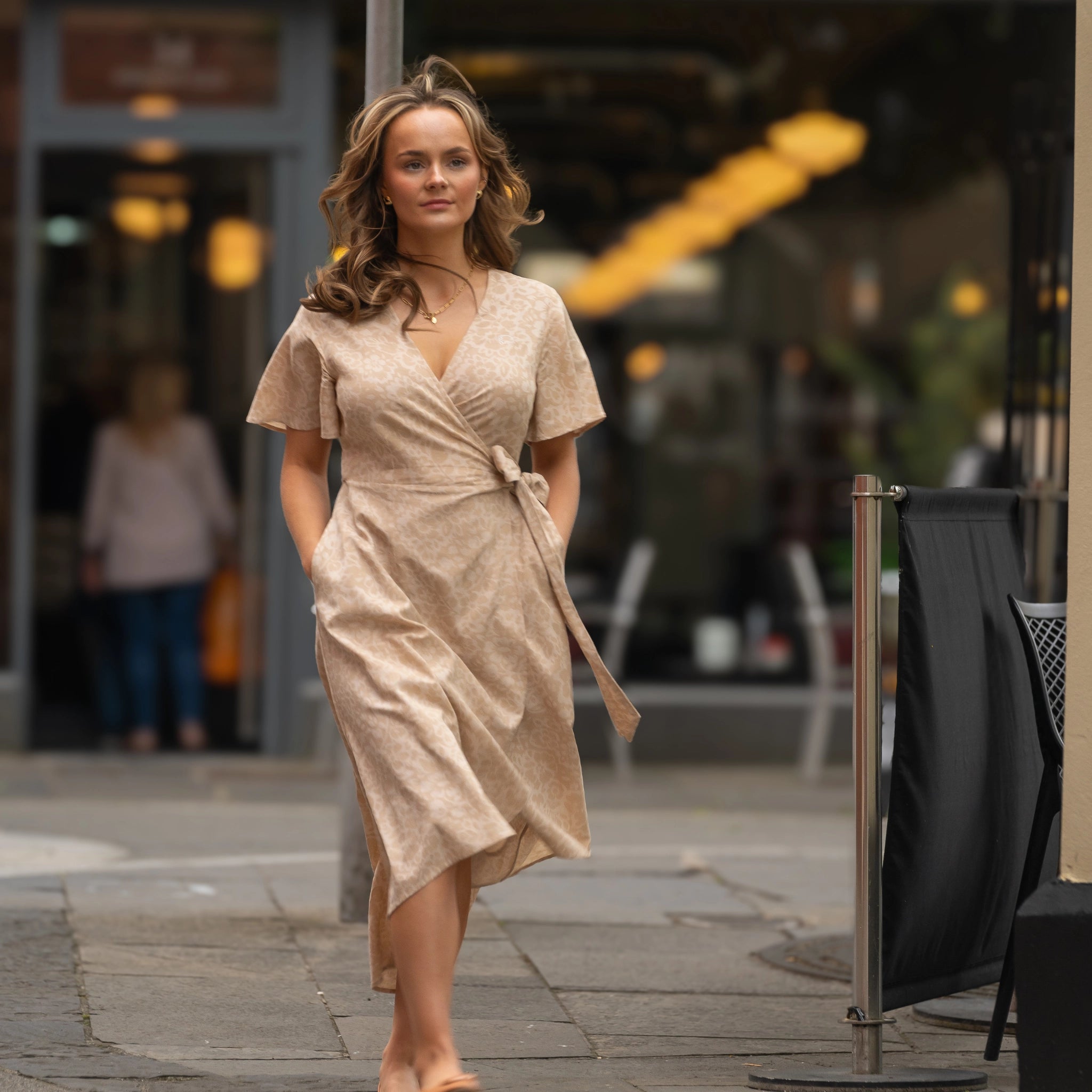 A woman in a beige wrap dress walks down a city street, with shops and people blurred in the background.