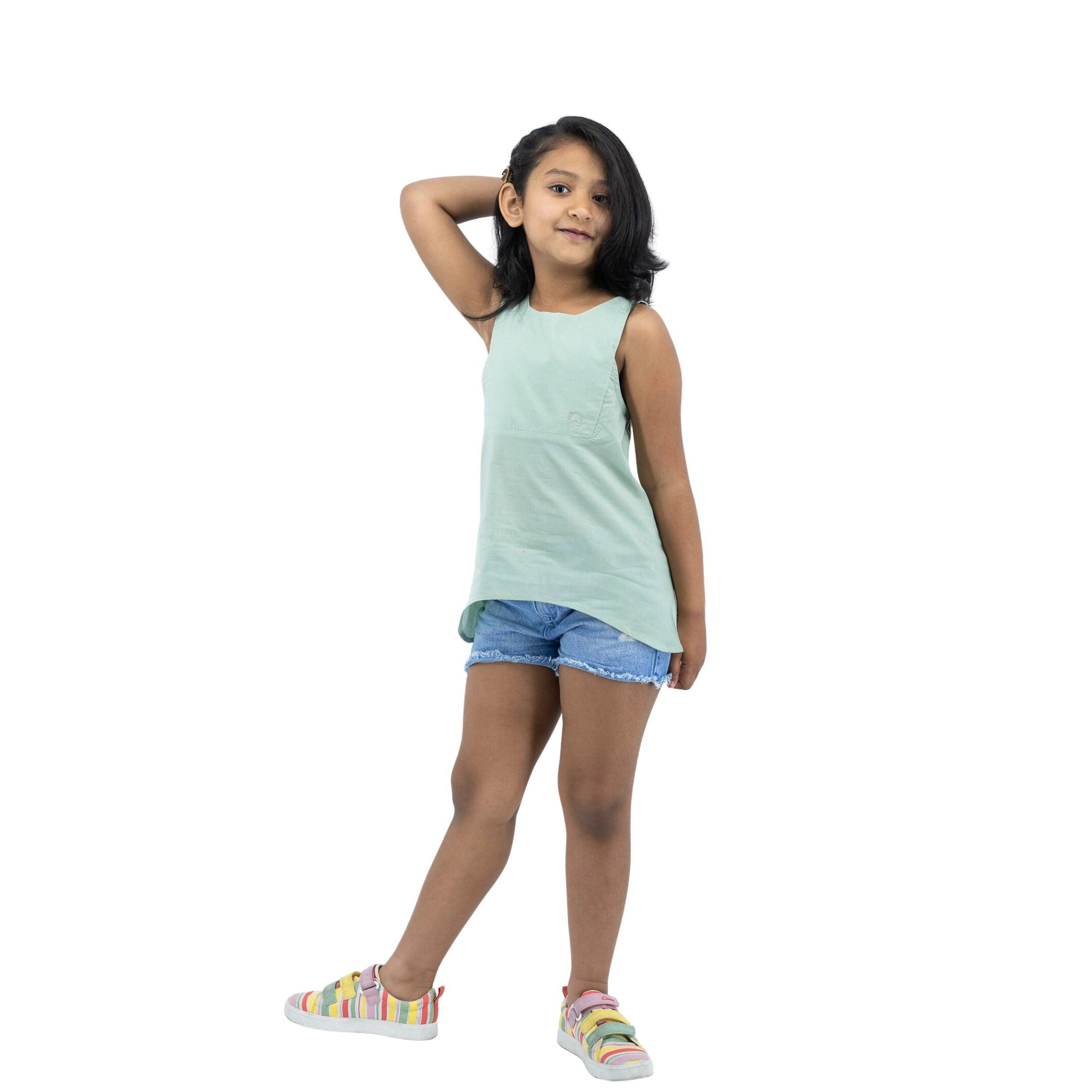 Young girl in a Smoke Green Cotton Bib Neck Top for kids by Karee and denim shorts, standing with one hand behind her head and smiling slightly, on a white background.