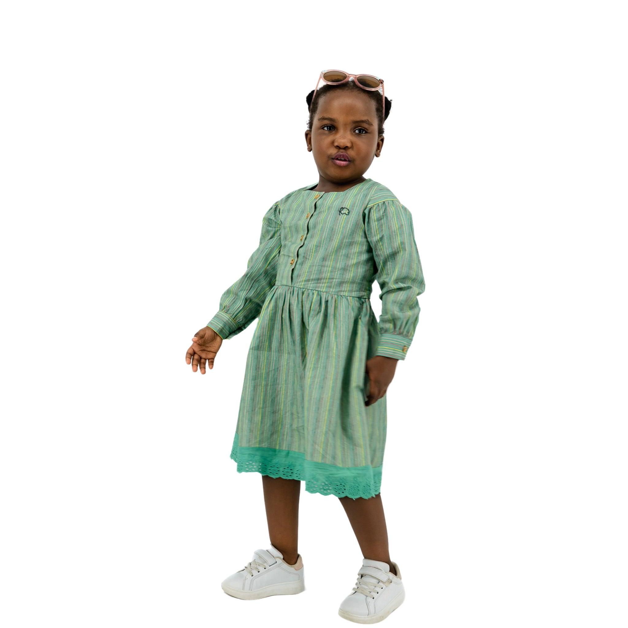 A young girl wearing a Karee Green Striped Long Puff Sleeve Cotton Dress and white sneakers, with sunglasses on her head, standing against a white background.