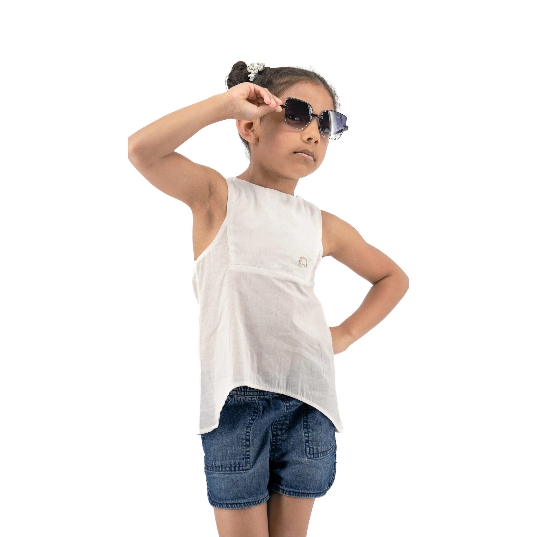 Young girl posing confidently in sunglasses, a Karee Brilliant White Cotton Bib Neck Top for kids, and denim shorts, with one hand adjusting her glasses.