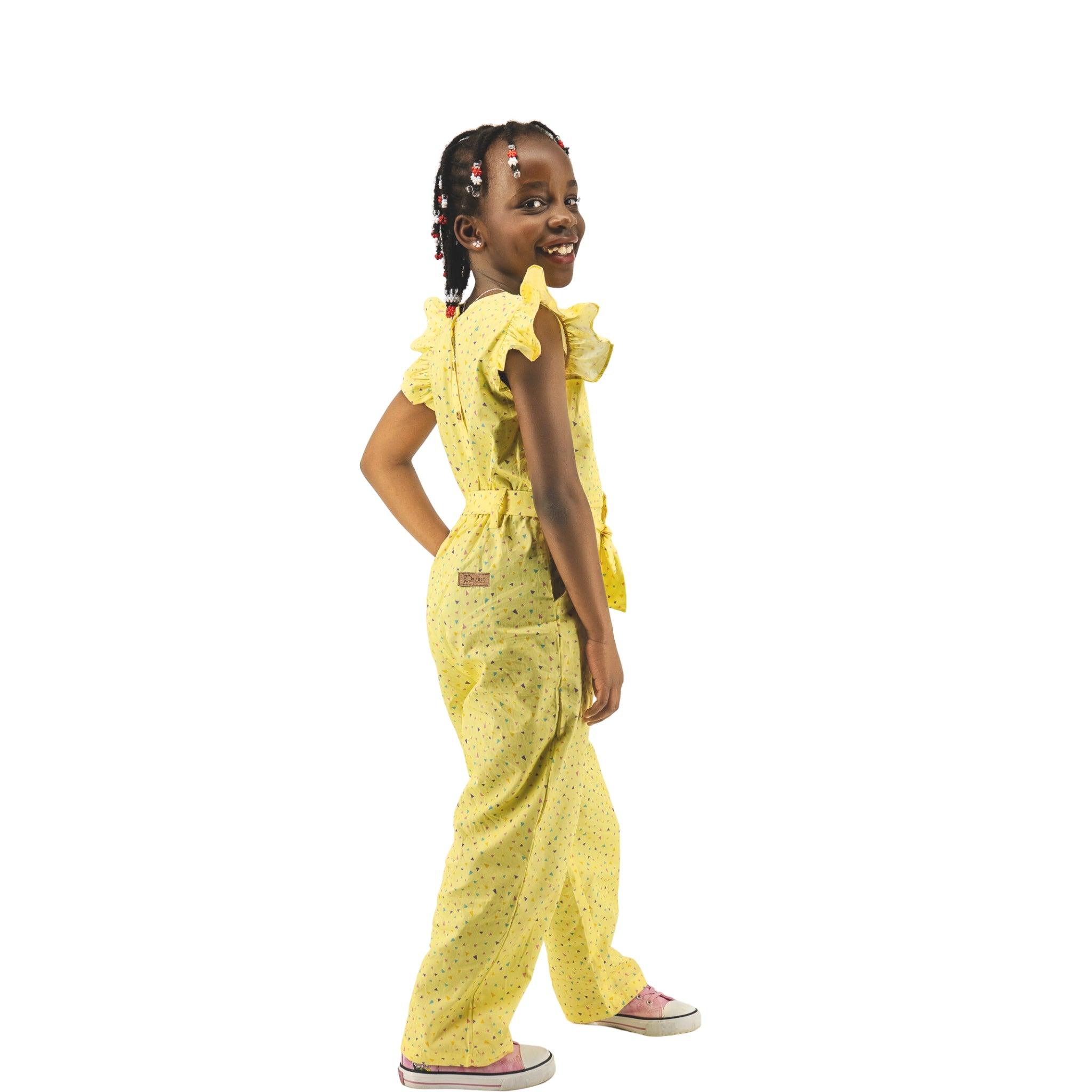 A young girl in a Karee Lemon Meringue Cotton V-Neck Jumpsuit smiling over her shoulder against a white background.