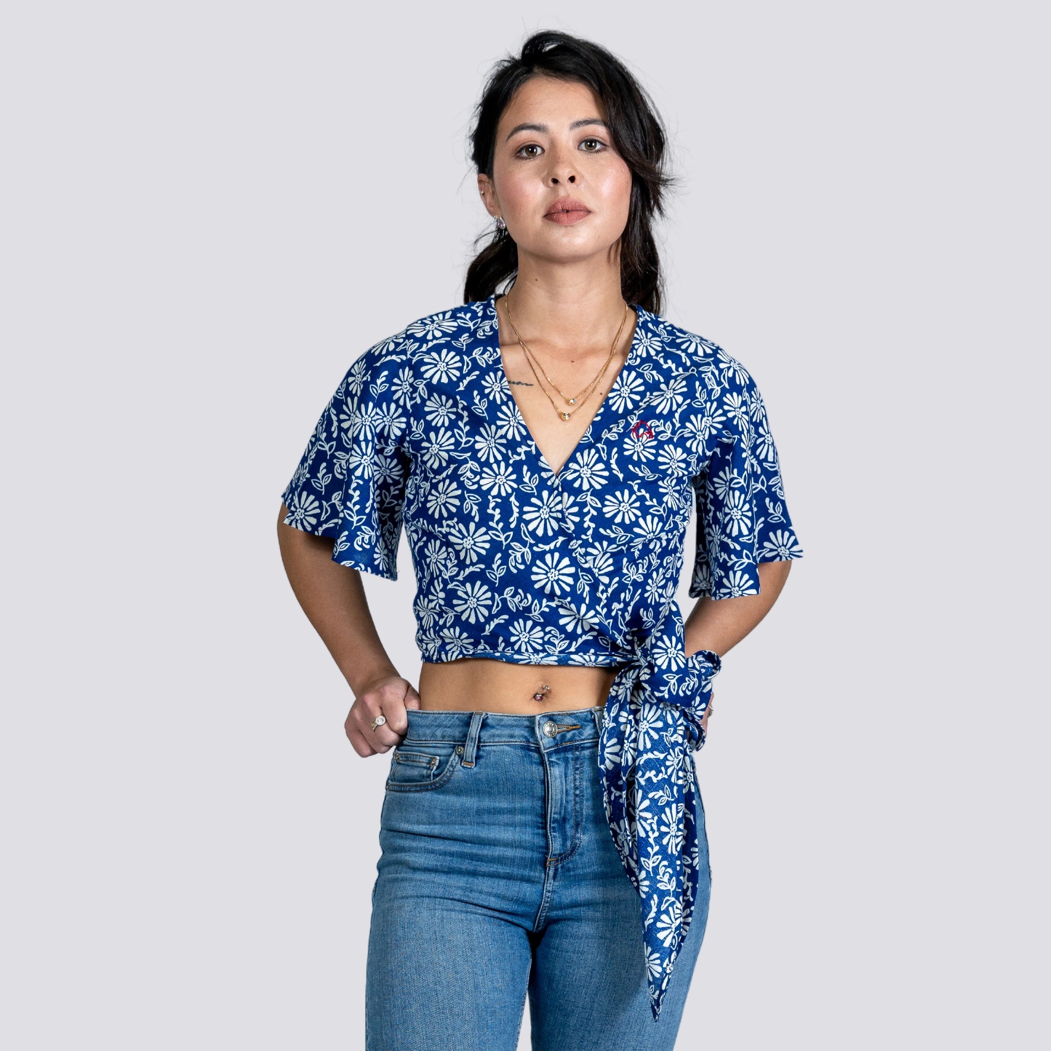 A young woman in a SereneBloom Women's Eco-Friendly Wrap Top - Blue Bliss by Karee and denim jeans stands confidently against a gray background.