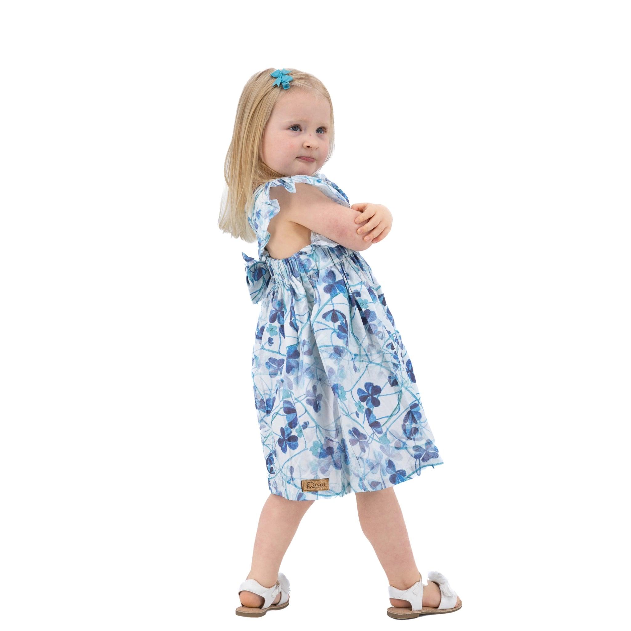 Toddler girl in a Karee blue cotton floral dress and white sandals standing sideways, looking over her shoulder, isolated on a white background.