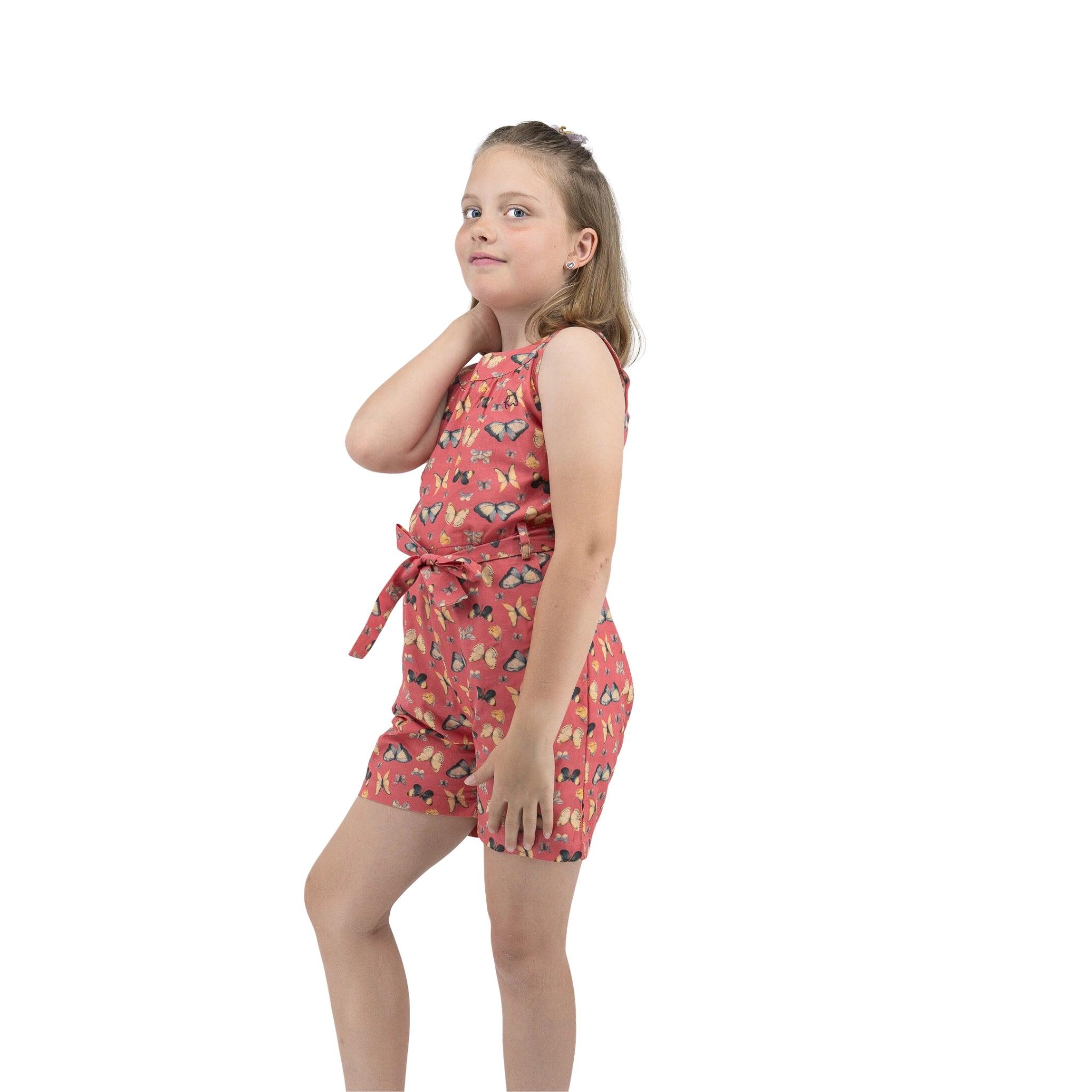 A young girl in a Karee Ruby Trek Cotton Romper posing with her hand on her neck, smiling slightly against a white background.