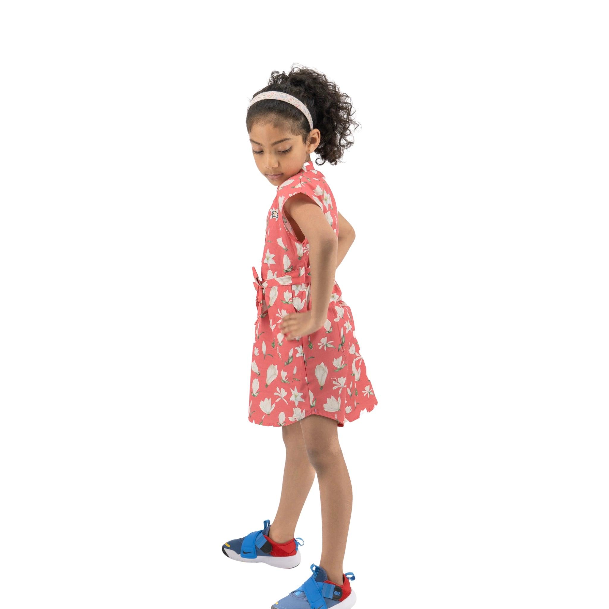 A young girl in a Karee Mineral Red Lilly Blossom Cotton Shirt Dress and blue shoes standing sideways, looking down, isolated on a white background.