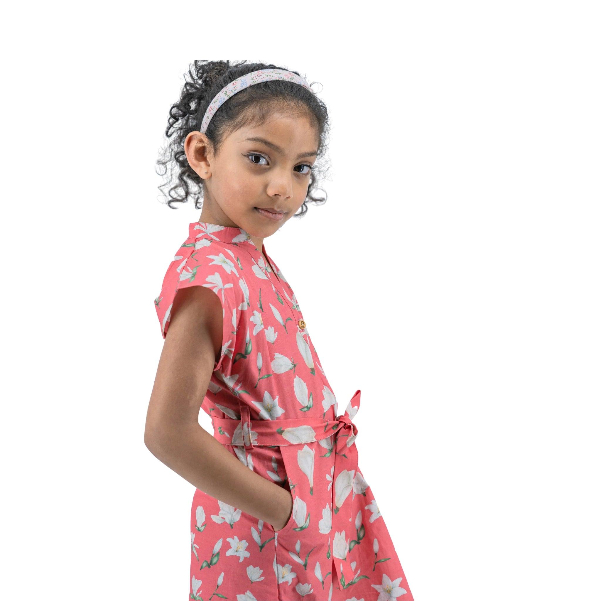 Young girl in a Karee Mineral Red Lilly Blossom Cotton Shirt Dress and headband standing sideways, looking over her shoulder at the camera, isolated on a white background.