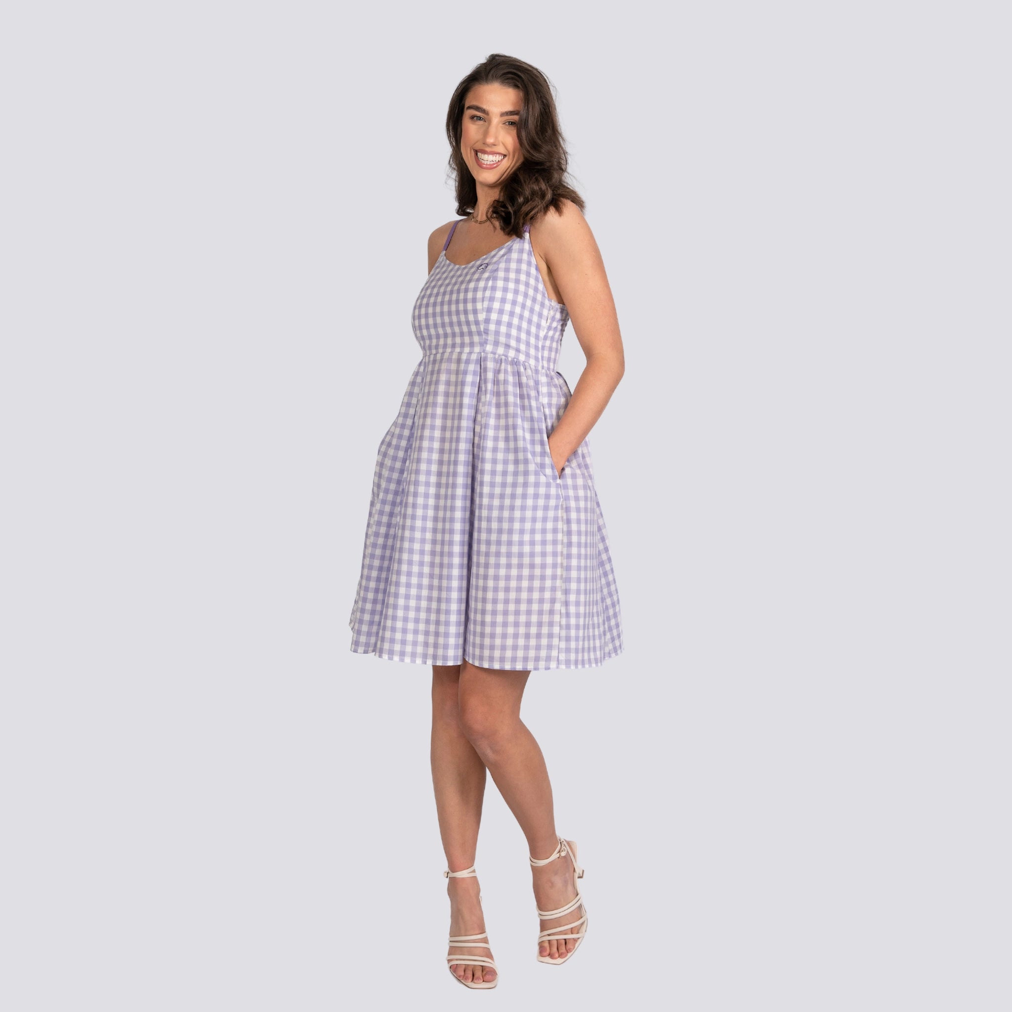 A woman in a Karee Effortless Summer Style: Sustainable Lavender Gingham Mini Dress stands smiling with hands in pockets, wearing white strappy heels against a plain light gray background.