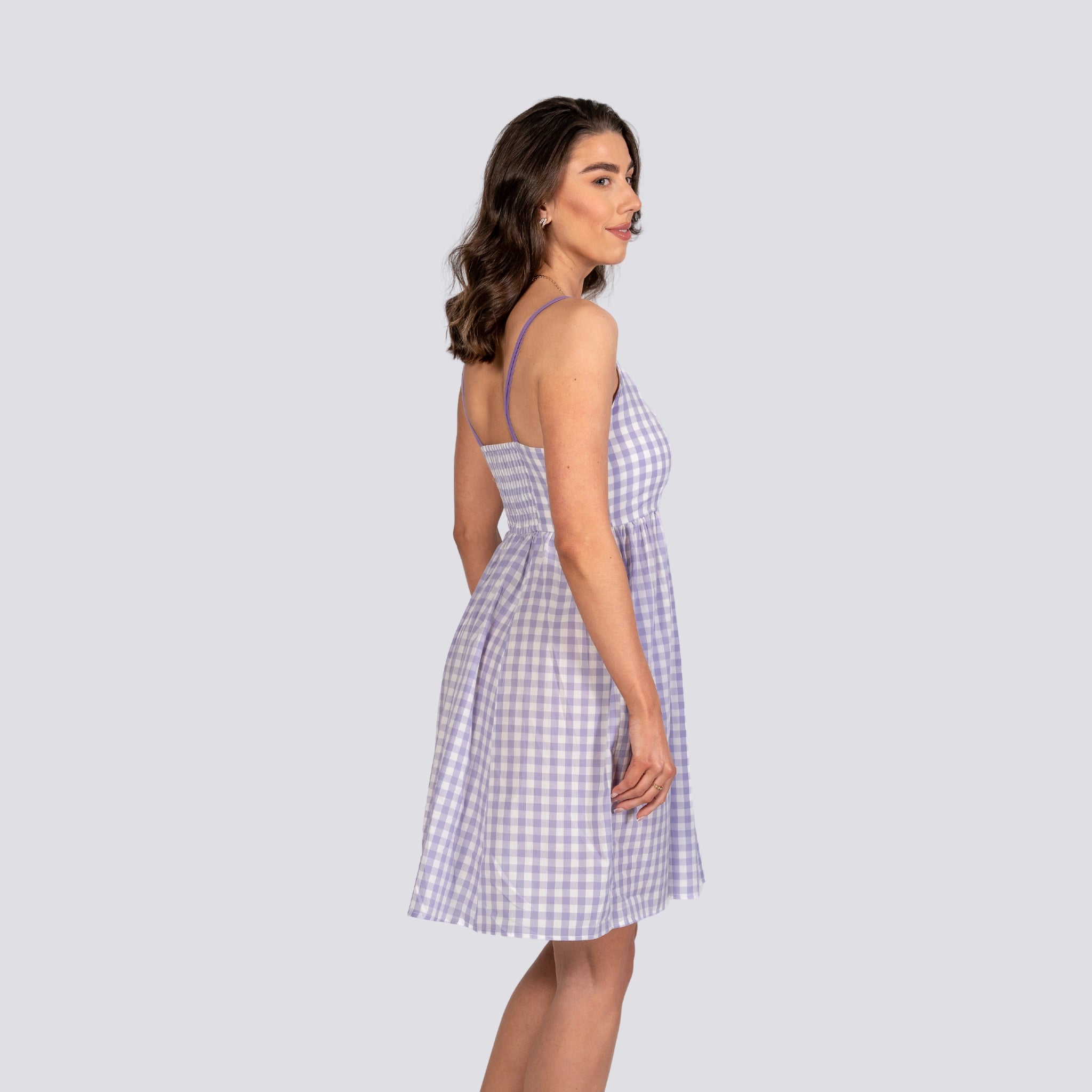 Woman in an Effortless Summer Style: Sustainable Lavender Gingham Mini Dress by Karee, standing sideways against a plain light gray background.