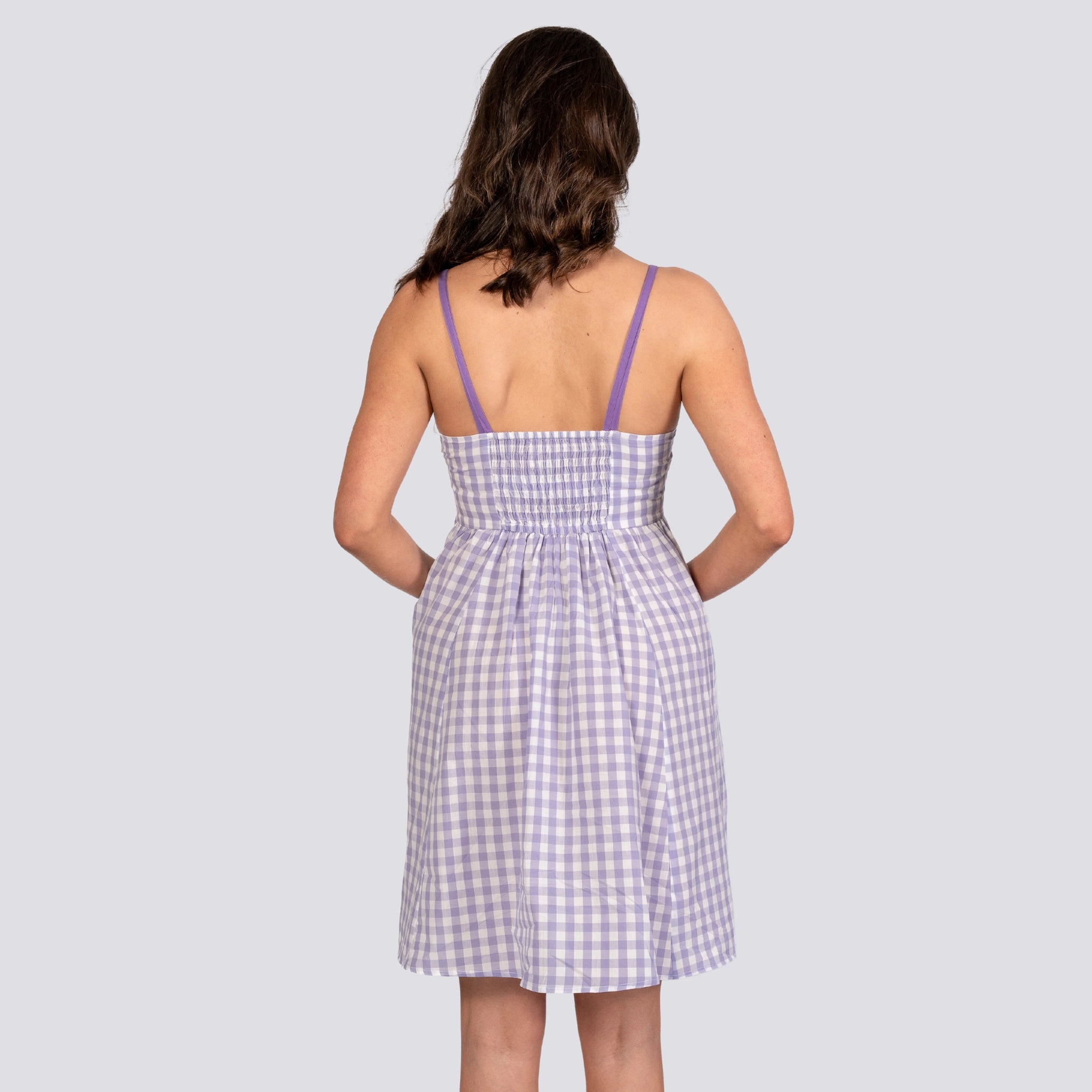 A woman with wavy brown hair is standing with her back to the camera, wearing a sleeveless Effortless Summer Style: Sustainable Lavender Gingham Mini Dress made from breathable cotton by Karee.