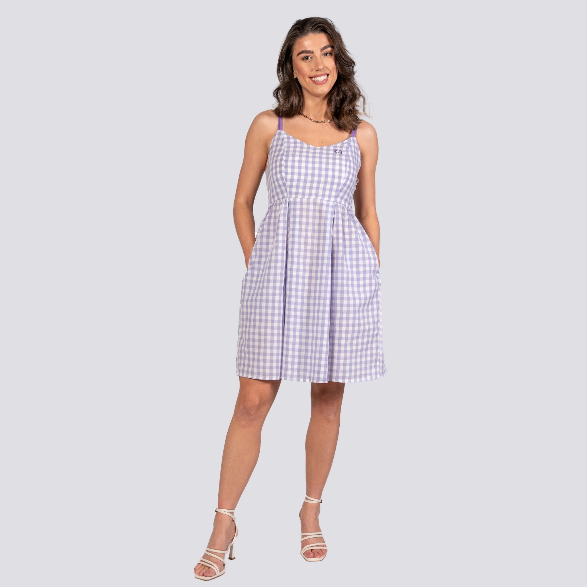 A woman in an Effortless Summer Style: Sustainable Lavender Gingham Mini Dress by Karee and white heels stands smiling against a plain background.